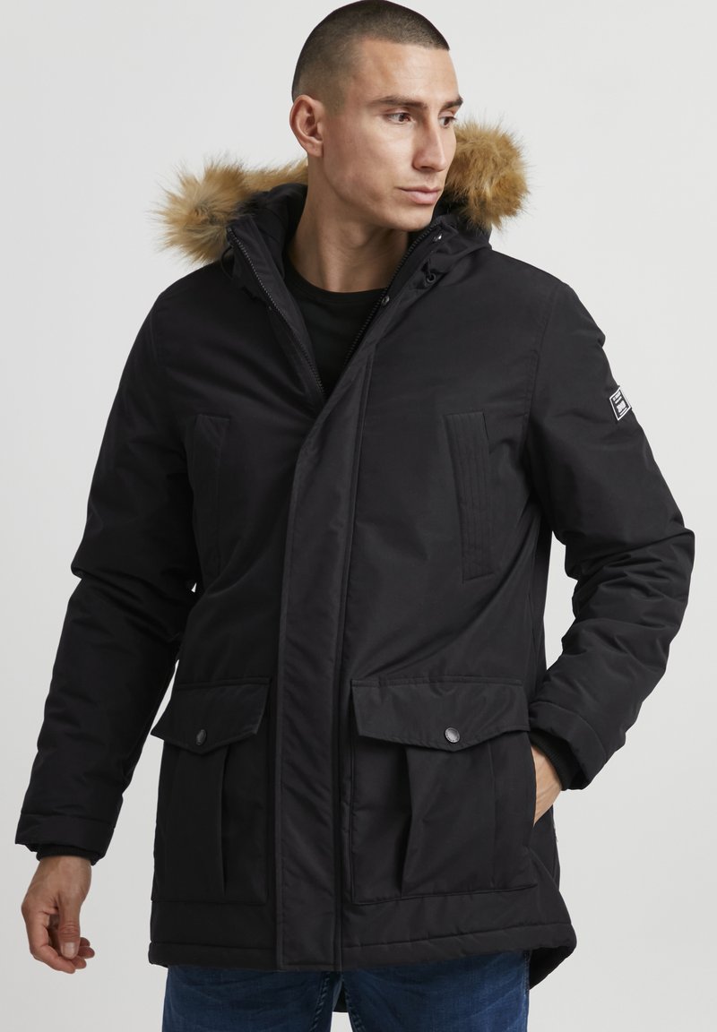 Sort polstret parka med pelsforet hætte, to forhåndsrum, lynlås og tekstureret detalje på brystet. Holdbart materiale, afslappet pasform.
