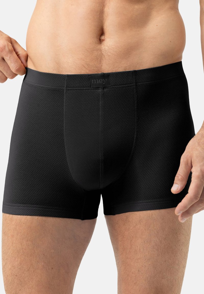 Buste masculin portant un boxer noir texturé, avec une main ajustant la taille, sur un fond blanc uni.