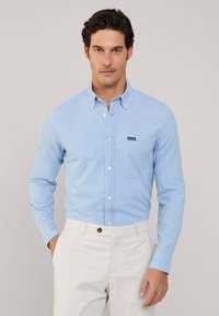 Camisa de botones a rayas azul claro hecha de un tejido de textura suave. Presenta un bolsillo de parche y un collar abotonado. Se lleva con pantalones de color claro.