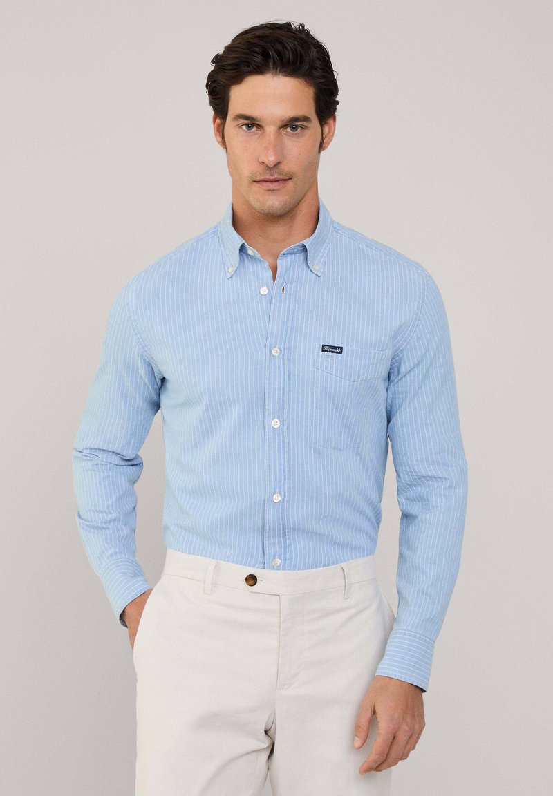 Camisa de botones a rayas azul claro hecha de un tejido de textura suave. Presenta un bolsillo de parche y un collar abotonado. Se lleva con pantalones de color claro.