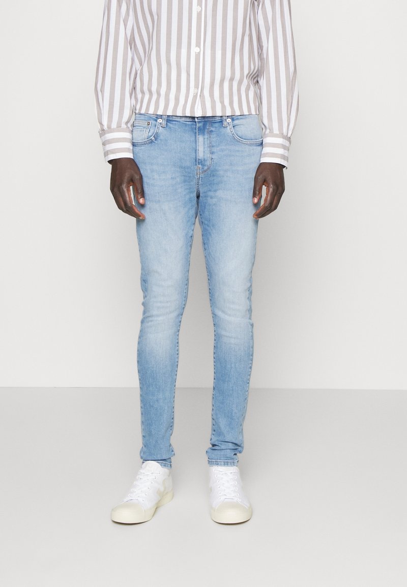 Hombre vistiendo jeans ajustados de color azul claro, zapatillas blancas y una camisa de botones a rayas verticales beige y blancas, de pie contra un fondo liso.