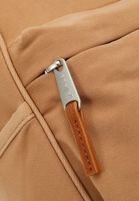 Tissu beige avec des coutures visibles et un tirette de fermeture éclair en argent gravé "aurelle", attaché à une languette en cuir marron.