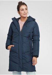 Jūras spēku puferskaids ar kapuci, ar quilted dizainu, ar rāvējslēdzēja priekšējām kabatām un ribotu piedurkņu apdari, uzvilkts virs krāsaina adīta džempera.
