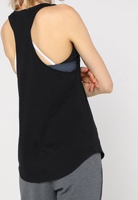 Svart ärmlös linne i bomull med rundad nederkant, stora armhål och racerback-design, lager på lager över en blå sportbh.