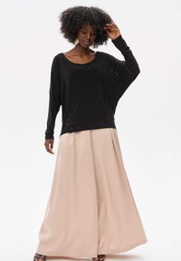 Schwarzes, weit geschnittenes Oberteil mit dezenten Verzierungen, kombiniert mit fließenden beigen, weit geschnittenen Hosen. Das Outfit zeichnet sich durch eine lässige Silhouette und weiche Texturen aus.