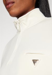 Witte ritsjack met hoge kraag, voorzien van een borstzak en een gouden driehoekembleem. Zachte textuur en een aansluitend ontwerp.