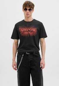 Musthave musta puuvillane T-särk, millel on punane graafika ja tekst "STRANGER THINGS". Sobib tumeate teksade ja vööle riputatud ahelaga.