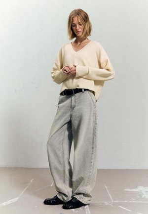 Kvinna med kort blont hår som bär en krämfärgad tröja, vida ljusgrå jeans, svart bälte och svarta skor, stående mot en plain vägg.