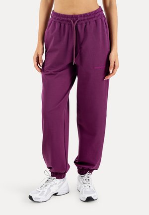 Kvinde iført lilla joggingbukser med elastiske manchetter og hvide atletiksneakers, stående mod en ensartet lys baggrund.