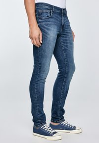 Mann trägt eng anliegende, ausgeblichene blaue Jeans und marineblaue Segeltuch-Sneaker mit weißen Schnürsenkeln, steht vor einem schlichten Hintergrund.