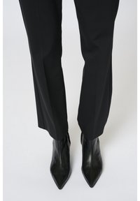 Personne portant des bottines en cuir noir à bout pointu associées à un pantalon noir droit et sur mesure, sur un fond uni.