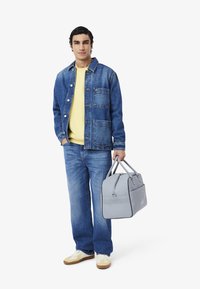 Spijkerjack met zakken over een gele trui, blauwe jeans en beige sneakers. Grijze duffelbag met geperforeerde accenten en ritssluiting.