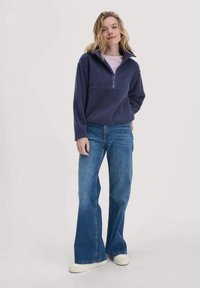 Marineblauer Fleece-Pullover mit halbem Reißverschluss und Stehkragen, kombiniert mit hochgeschnittenen blauen Jeans und weißen Sneakers. Einfaches, lässiges Design.