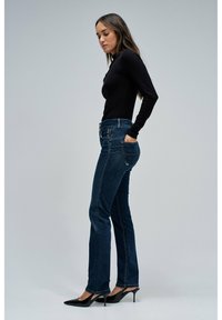 Jeans acampanados de mezclilla azul oscuro con cintura alta, con detalles de costura en el bolsillo trasero y combinados con tacones negros de punta afilada.