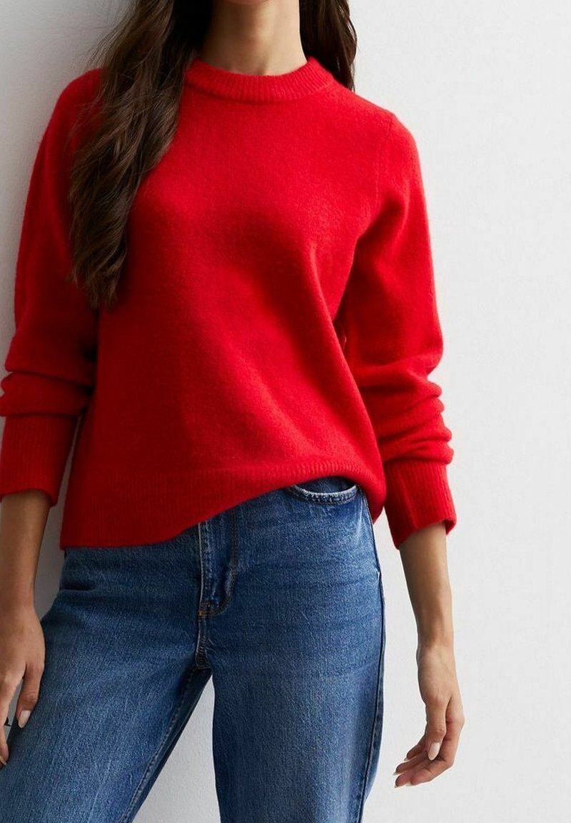 Femme portant un pull rouge vif à manches longues et un jean en denim bleu, debout devant un fond clair uni.