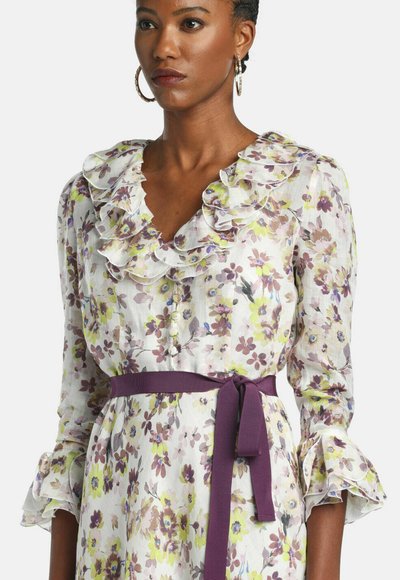 Femeie purtând o rochie albă cu flori galbene și mov, cu guler și manșete cu volane și o centură mov legată în talie.