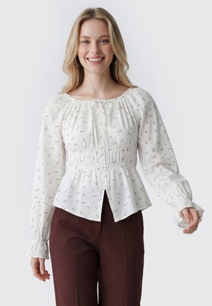 Lächelnde Frau mit blondem Haar trägt eine weiße langärmelige Bluse mit kleinem Blumenmuster und dunkelbraune Hose, steht vor einfarbigem Hintergrund.