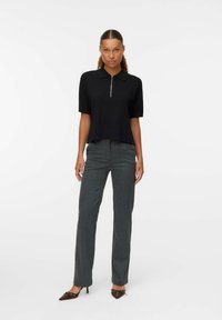 Polo noir à manches courtes avec une fermeture éclair devant, associé à un pantalon à motifs gris et des escarpins à bout pointu marron.