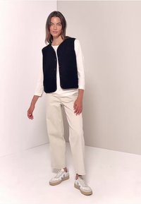 Svart, ärmlös väst med knappar, bärs över en vit långärmad skjorta. Matchas med ljusbeige vida byxor och vita sneakers.