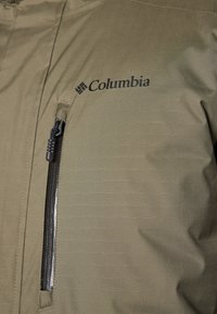 Olivgrüne Jacke mit strukturiertem Finish, ausgestattet mit einer Reißverschlusstasche und einem dunkelbedruckten Columbia-Logo auf der Brust.