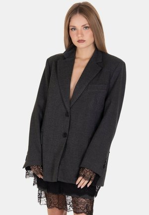 Blazer oversized di un grigio scuro, in tessuto texturizzato, con chiusura monopetto, rever a punta e dettagli in bottone. Accenti di pizzo che spuntano da sotto.