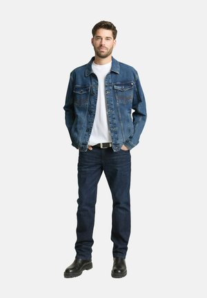 Mann steht mit den Händen in den Taschen, trägt eine blaue Jeansjacke, ein weißes Hemd, dunkle Jeans und schwarze Stiefel vor weißem Hintergrund.