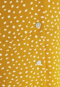 Senfgelber Stoff mit verstreuten weißen abstrakten Formen, ausgestattet mit einem Knopfverschluss aus glänzendem Material, glatte Textur.