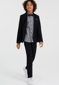 Svart kavaj med texturerad tyg, grå skjorta med mönster, svarta byxor och vita sneakers; fluga som accent.