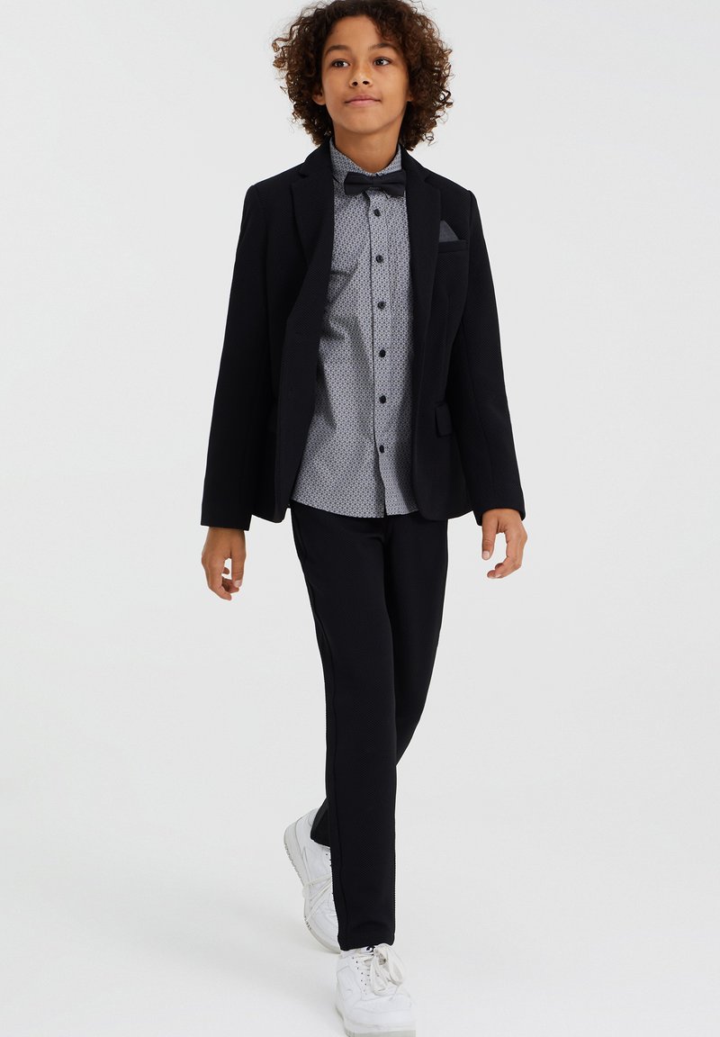 Svart kavaj med texturerad tyg, grå skjorta med mönster, svarta byxor och vita sneakers; fluga som accent.