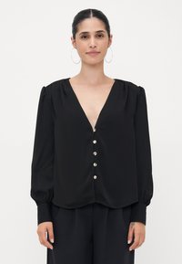 La blusa nera presenta maniche lunghe, un profondo scollo a V, spalle plissettate e dettagli con bottoni decorativi lungo il davanti. Tessuto dalla trama liscia.