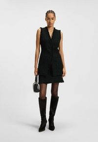 Schwarzes Tweed ärmelloses Vestkleid mit geradem Schnitt, Knopfleiste und fransigen Kanten, kombiniert mit kniehohen schwarzen Wildlederstiefeln.