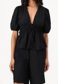 Blusa peplo nera con maniche corte a sbuffo, scollatura a V profonda e vita regolabile con laccetto. Tessuto morbido, leggero e vestibilità rilassata.