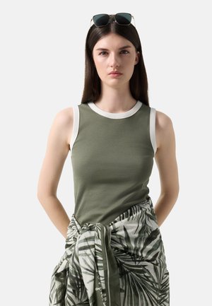 Jonge vrouw met stijlvol donker haar, gekleed in een groene mouwloze top met witte rand, een zonnebril op haar hoofd en een stof met patroon om haar middel geknoopt.