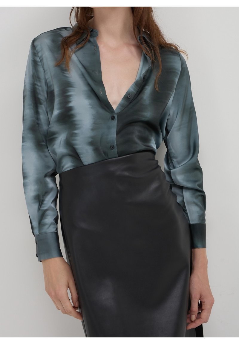 Femme portant un chemisier tie-dye gris-bleu brillant, rentré dans une jupe en cuir noire taille haute, debout devant un fond uni.