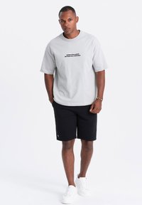 T-shirt en coton gris clair à manches courtes avec texte noir. Porté avec des shorts noirs texturés et des baskets blanches. Tenue décontractée.