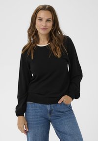 Kvinde med bølget brunt hår if�ørt guldøreringe, en sort langærmet sweater og blå jeans, stående med den ene hånd i lommen.