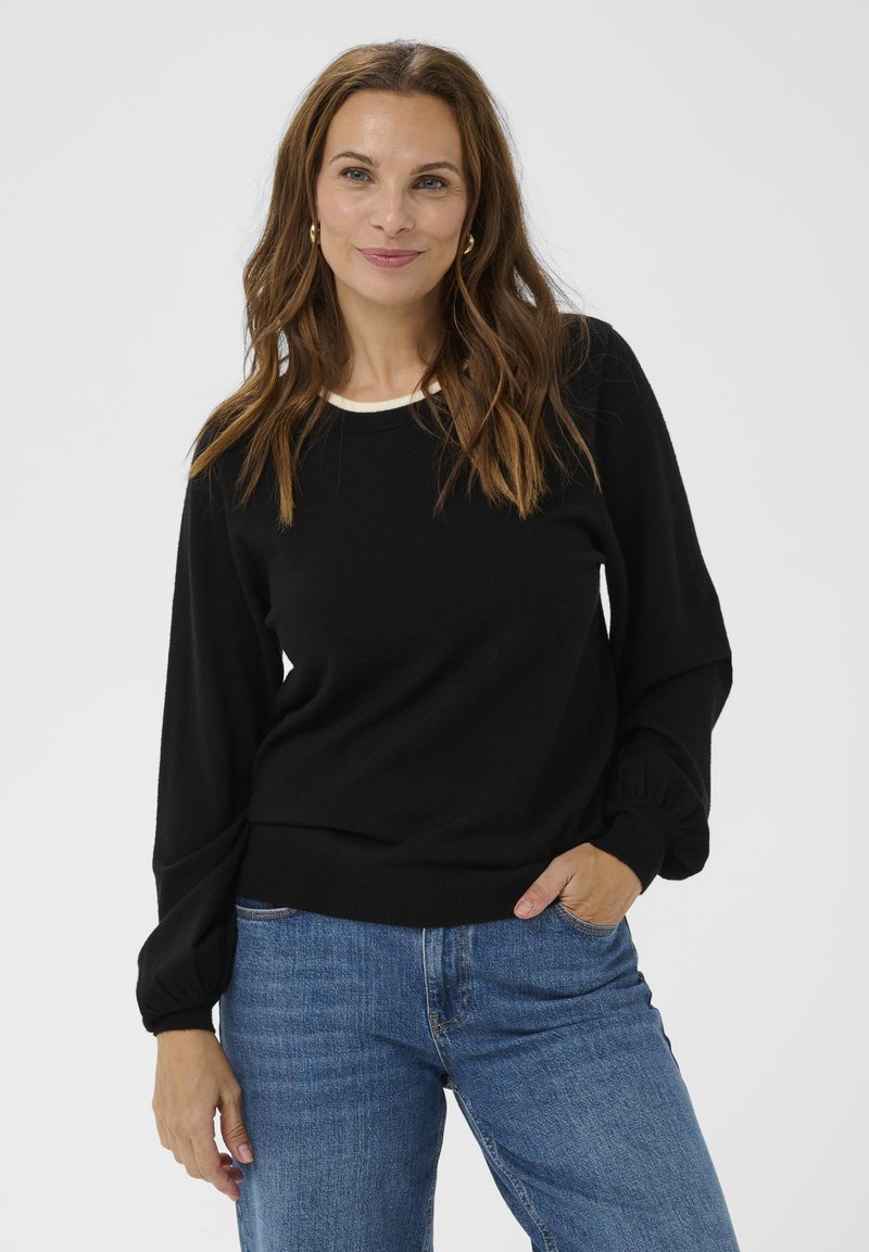 Kvinde med bølget brunt hår iført guldøreringe, en sort langærmet sweater og blå jeans, stående med den ene hånd i lommen.