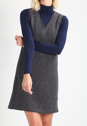 Person wearing a sleeveless gray wool dress over a navy blue turtleneck with arms crossed in front against a plain light background.