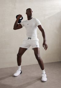Camiseta y pantalones cortos deportivos Nike blancos, sosteniendo un kettlebell negro. Las características incluyen un diseño ajustado, acentos de logo y calzado deportivo.