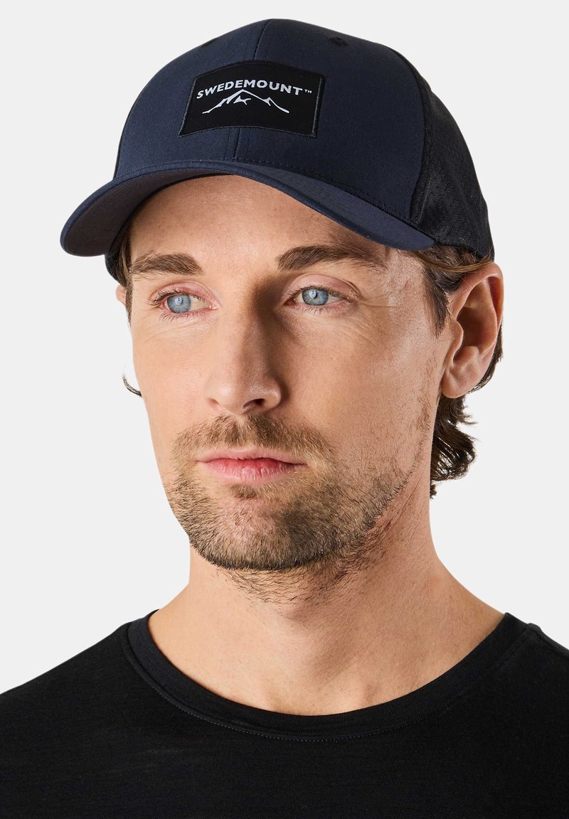 Homme avec une légère barbe portant une casquette bleue marine Swedemount et un t-shirt noir à col rond, regardant légèrement sur le côté sur un fond blanc.