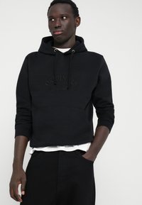 Sweatshirt noir à capuche avec logo en relief "ORIGINALS", poche kangourou et bords côtes. Porté sur un t-shirt blanc et associé à un pantalon noir.