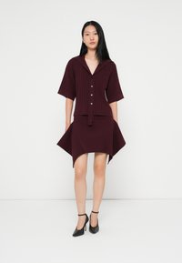Vestido bordeaux com textura canelada, mangas curtas, fecho de botões e bainha assimétrica. Combinado com sapatos de salto alto pretos.