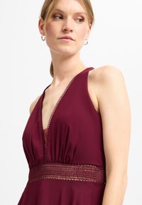 Burgunderrotes Kleid mit tiefem V-Ausschnitt, gekennzeichnet durch gerafftes Gewebe und Häkelverzierungen an der Taille, aus glattem Material gefertigt.