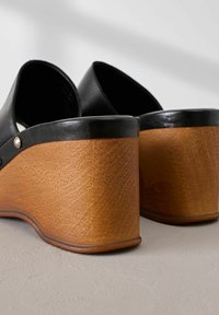 Sandali con zeppa in pelle nera, dotati di un tacco in legno marrone texturizzato, con un retro arrotondato e dettagli metallici sui lati.