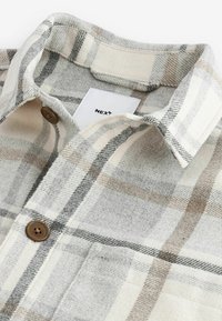 Gros plan sur le col d’une chemise à carreaux beige et gris avec des boutons marron et une étiquette de marque blanche visible à l’intérieur du col.