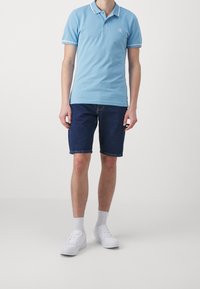 Jasnoniebieska koszulka polo z białymi akcentami, zestawiona z ciemnymi szortami dżinsowymi. Model nosi białe trampki i skarpetki, stojąc na jednolitym tle.