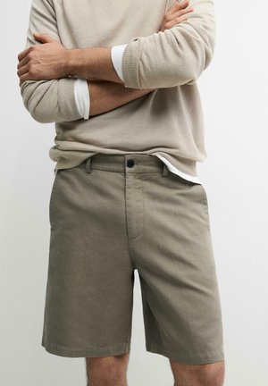 Mann trägt einen beigen Pullover mit langen Ärmeln über einem weißen Hemd und olivgrüne knielange Shorts, die Arme vor weißem, einfarbigem Hintergrund verschränkt.