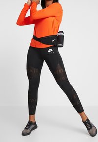 Zwarte Nike-legging met gaasdetails, gecombineerd met een oranje top met lange mouwen en een zwarte heuptas met een waterfles.