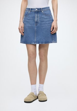 Femme portant une jupe en jean bleu coupe trapèze, des chaussettes blanches, des chaussures beiges à enfiler et un haut blanc, debout devant un fond uni.