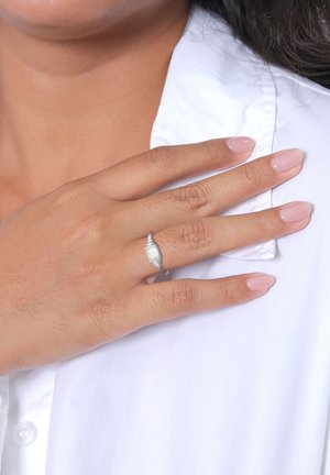 Silberner Ring mit filigranem Design, mit rechteckiger Steinfassung, auf einer Hand vor einem weißen Hemd-Hintergrund dargestellt.